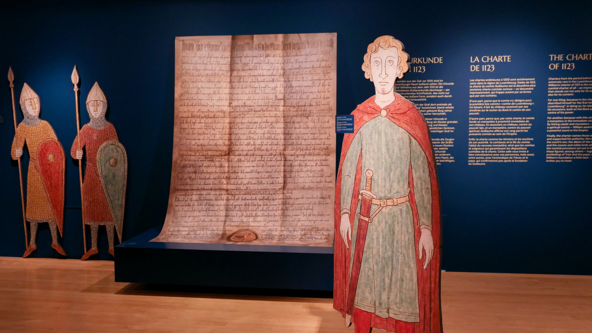 Exhibition room with an enlarged reproduction of the 1123 charter, flanked by painted cut-out figures of two knights with spears and shields and Count William in a red cloak
